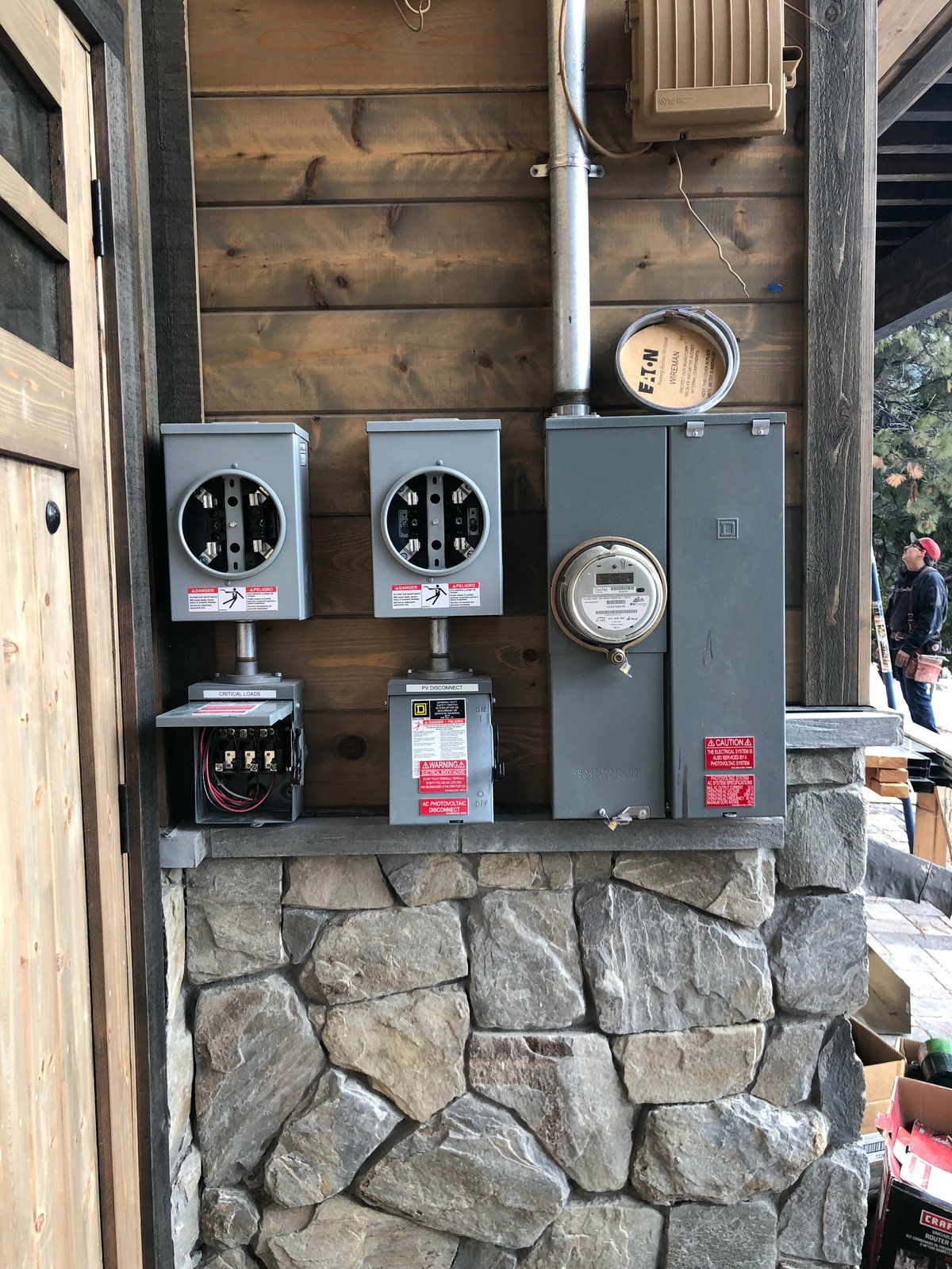 Exterior electrical panel mounted on stone base with two analog meters and circuit boxes on wooden house wall